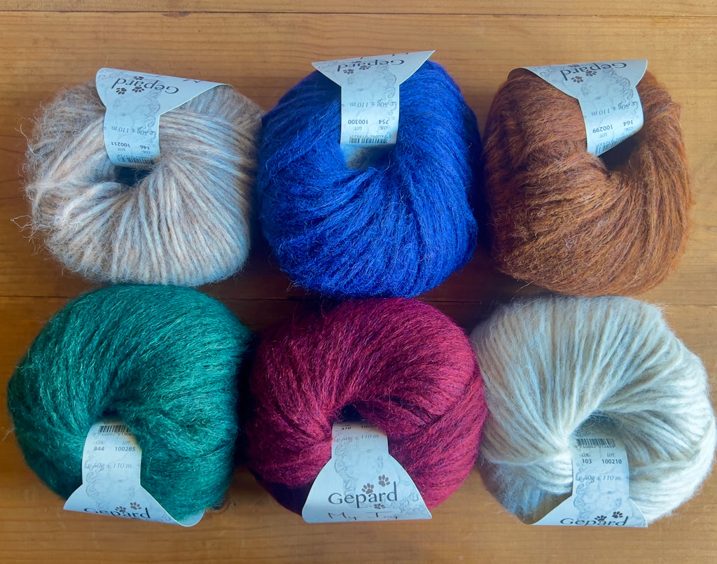 Six balls of Gepard My Joy yarn in taupe, blue, copper brown, green, burgundy and off-white, arranged on a wooden surface